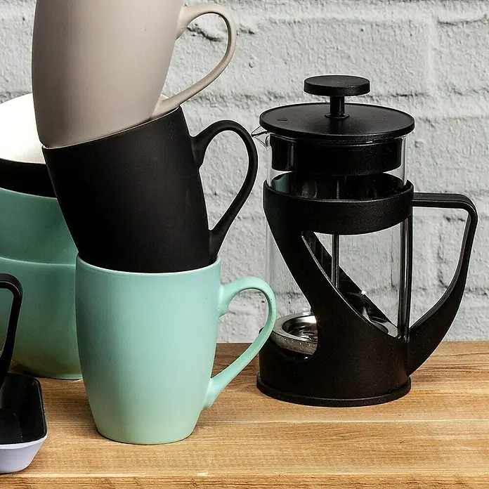 Cafetera de émbolo - 600 ml, 5 tazas - Una pila de tazas de colores junto a una elegante cafetera negra sobre una mesa de madera frente a una pared de ladrillo blanco.