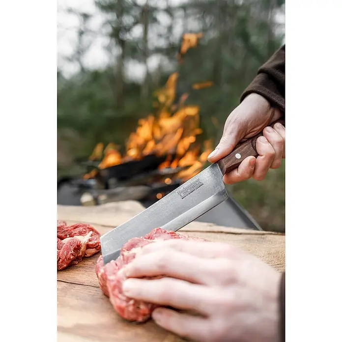 Petromax Küchenmesser - Stahl, 31,5 cm - Ein Petromax Hackmesser im Einsatz, das auf einem Holzbrett Fleisch schneidet, mit einem Lagerfeuer im Hintergrund.