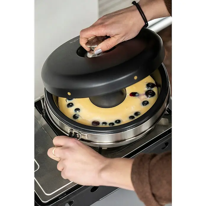 Petromax Backofenaufsatz Dimego - Ø x H: 27 x 10 cm - Der Omnia Backofen in Aktion: Eine Person hebt den Deckel über einem Kuchen mit Blaubeeren, der auf einem Gaskocher steht.