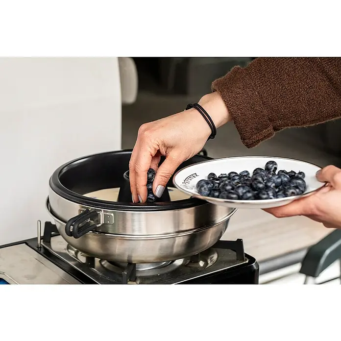 Petromax Backofenaufsatz Dimego - Ø x H: 27 x 10 cm - Eine Person fügt Blaubeeren zu einem Kuchen hinzu, der in einer Petromax Kastenform auf einem Herd gebacken wird.