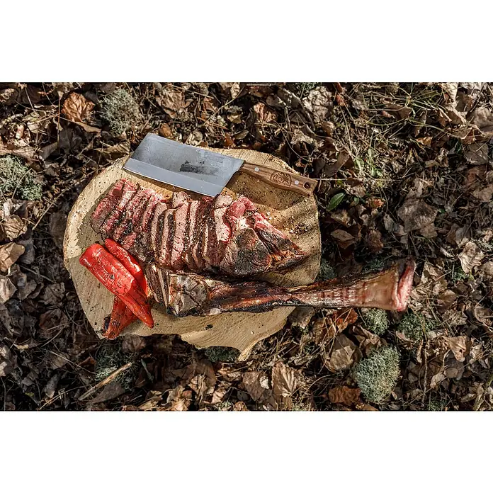Petromax Hackmesser - Stahl, Länge: 32,5 cm - Ein saftiges Steak mit Knochen, serviert mit gegrillter roter Paprika und einem Hackmesser auf einem Holzhocker im Freien.