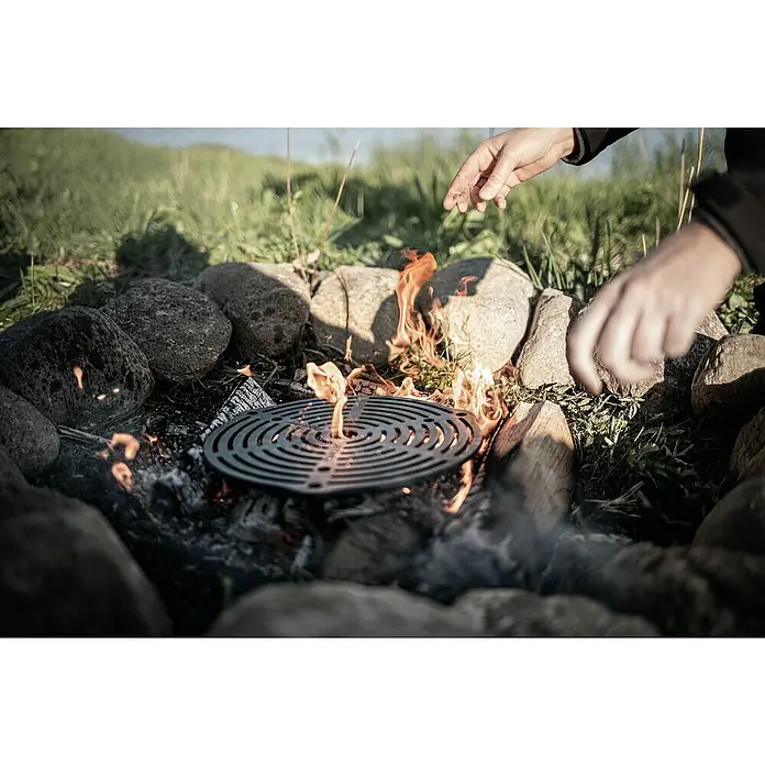 Petromax Grillrost zum Stapeln aus Gusseisen - Durchmesser: 30 cm, Gusseisen - Ein Lagerfeuer mit einem runden Grillrost, umgeben von Steinen und lodernden Flammen, ideal zum Kochen im Freien.