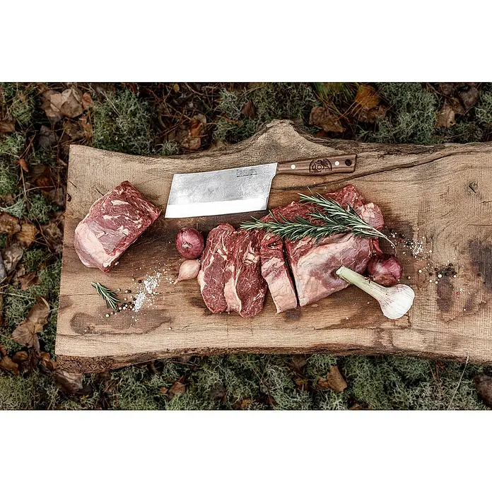 Petromax Hackmesser - Stahl, Länge: 32,5 cm - Auf einem rustikalen Holzbrett liegt rohes Rindfleisch, ein Hackmesser, Knoblauch, Zwiebeln und Gewürze, bereit für die Zubereitung.