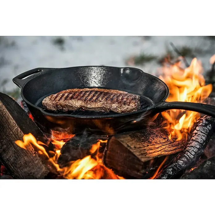 Petromax Grillpfanne - Durchmesser: 30 cm, Höhe: 4,8 cm, Gusseisen - Ein saftiges Steak brutzelt in einer schwarzen gusseisernen Pfanne über einem Lagerfeuer. Köstliche Röstaromen entstehen beim Grillen.