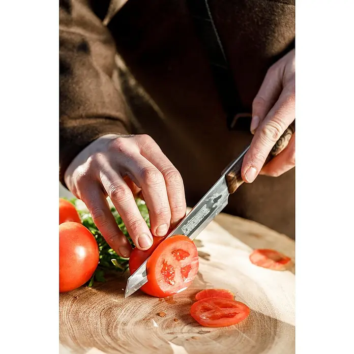 Petromax Allzweckmesser - Stahl, Länge: 28 cm - Nahaufnahme einer Person, die mit einem scharfen Messer eine rote Tomate auf einem Holzbrett schneidet. Perfekt für kulinarische Zwecke.