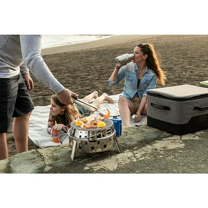 Petromax Gasgrill Atago - Durchmesser Grillfläche: 28 cm, Edelstahl - Ein tragbarer Grill von Feuerdesign im Einsatz am Strand, mit Familie im Hintergrund. Ideal für ein Picknick mit leckerem Essen.