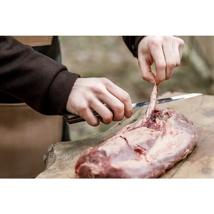 Petromax Allzweckmesser - Stahl, Länge: 28 cm - Nahaufnahme einer Person, die mit einem Messer ein Stück rohes Fleisch auf einem Holzbrett bearbeitet, wobei die Textur und Frische des Fleisches betont werden.