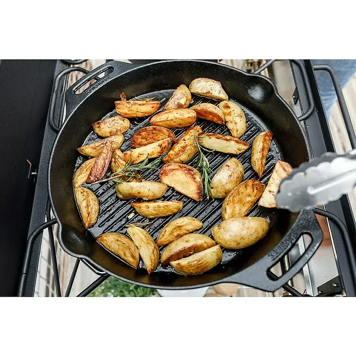Petromax Grillpfanne - Durchmesser: 35 cm, Höhe: 5 cm, Gusseisen - Kartoffelspalten werden in einer gusseisernen Grillpfanne mit Rosmarin zubereitet. Eine Zange hält eine Kartoffel über der Pfanne.