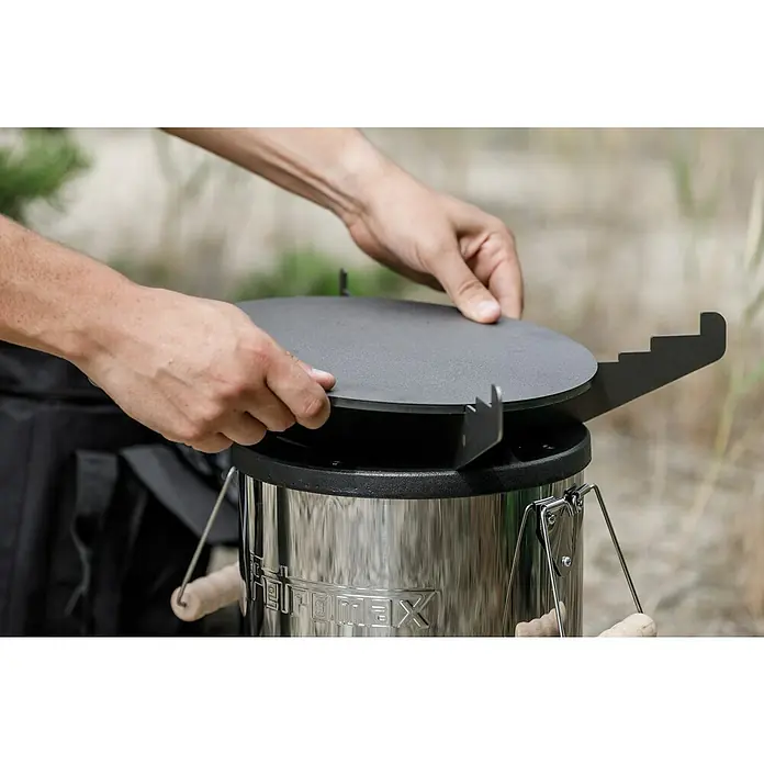 Petromax Grillplatte - Durchmesser: 27,5 cm, Passend für: Petromax Raketenofen - Nahaufnahme eines Grills mit einer Person, die einen Deckel aufsetzt. Der Deckel ist rund und grau.
