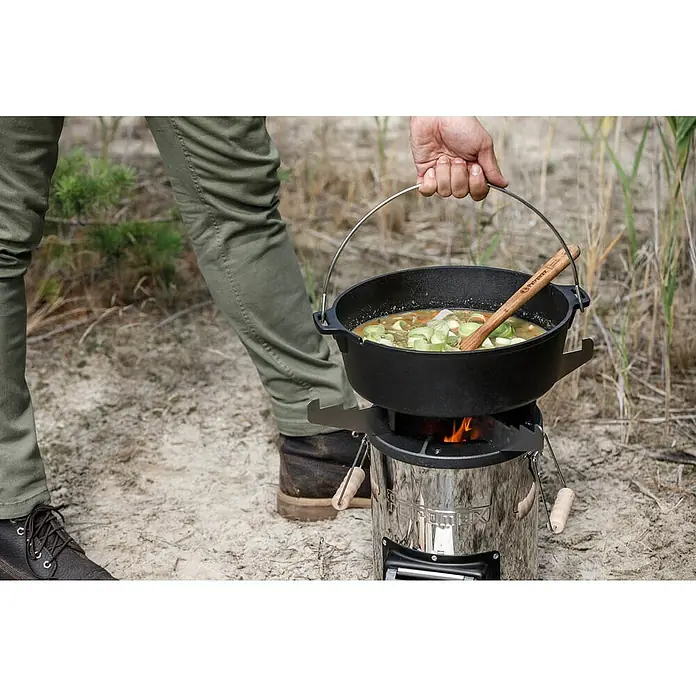 Petromax Grillplatte - Durchmesser: 27,5 cm, Passend für: Petromax Raketenofen - Ein Mann kocht eine Mahlzeit im Freien mit einem Petromax Feuertopf auf einem robusten Kocher, umgeben von natürlicher Umgebung.
