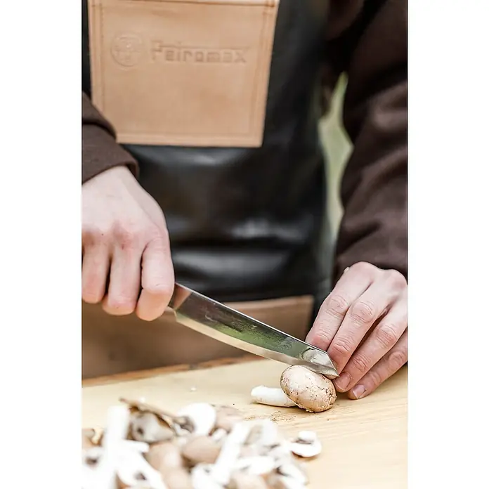 Petromax Allzweckmesser - Stahl, Länge: 28 cm - Eine Person, die mit einem Messer auf einem Holzbrett Champignons schneidet, trägt dabei eine Petromax Lederschürze.