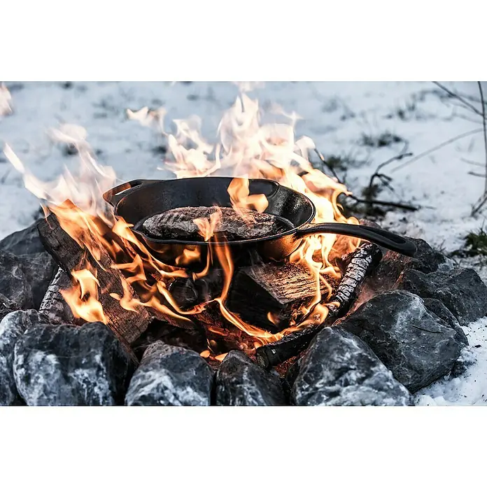Petromax Grillpfanne - Durchmesser: 30 cm, Höhe: 4,8 cm, Gusseisen - Ein Steak brutzelt in einer gusseisernen Pfanne über einem Lagerfeuer, umgeben von Steinen und Schnee. Köstliche Outdoor-Küche.