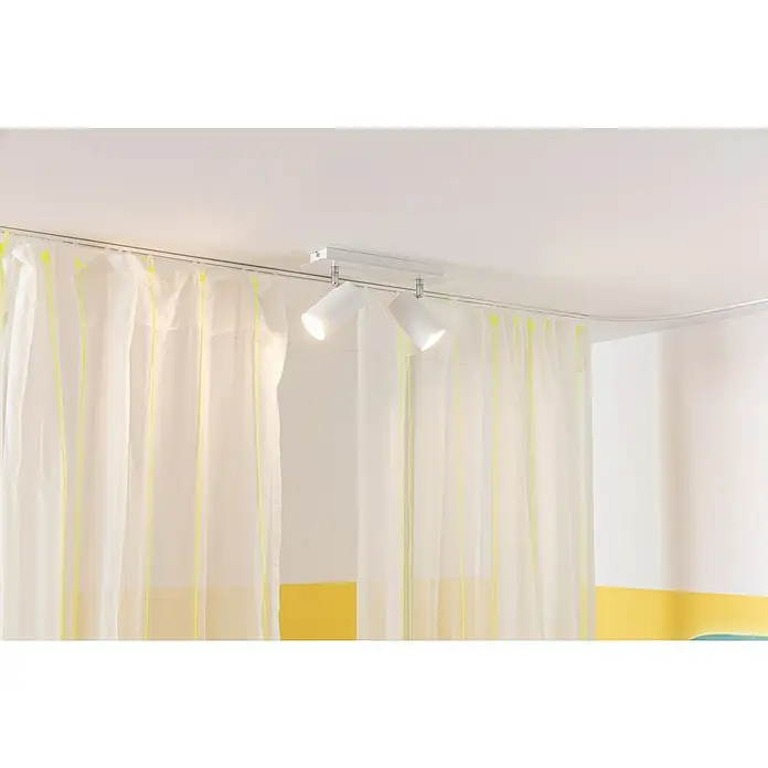 Elbersdrucke Vorhang Spume - Offwhite, 255 x 200 cm, 100% Polyester - Nahaufnahme einer modernen Innenraumbeleuchtung mit weißen Strahlern an einer Schiene unterhalb der Decke. Dazu helle Vorhänge mit gelben Akzenten.