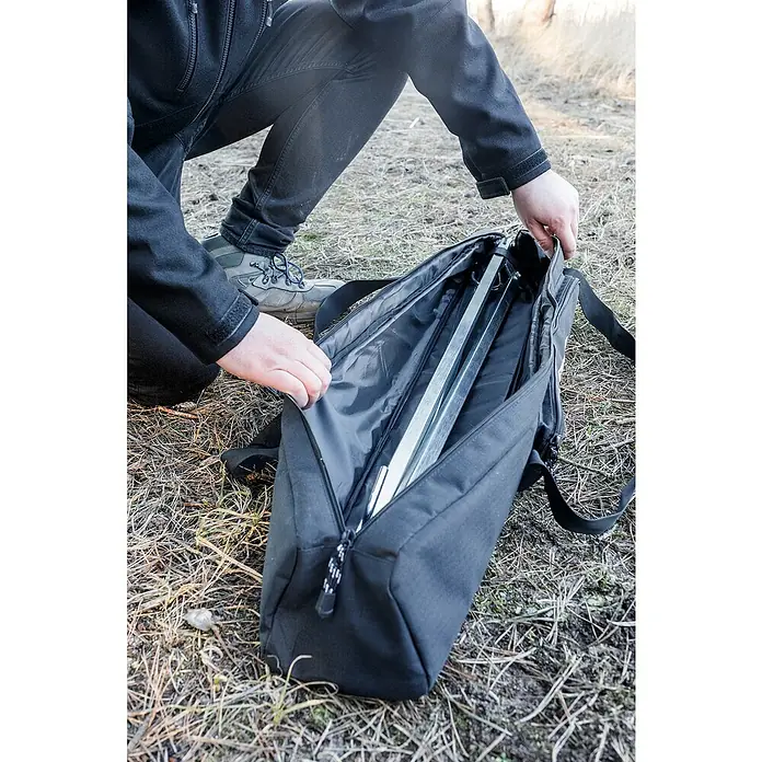 Petromax Transporttasche Dreibeinständer - Nylon, Passend für: Petromax Dreibeinständer - Eine Person kniet und öffnet eine schwarze Tragetasche mit Reißverschluss, in der sich ein Stativ befindet.