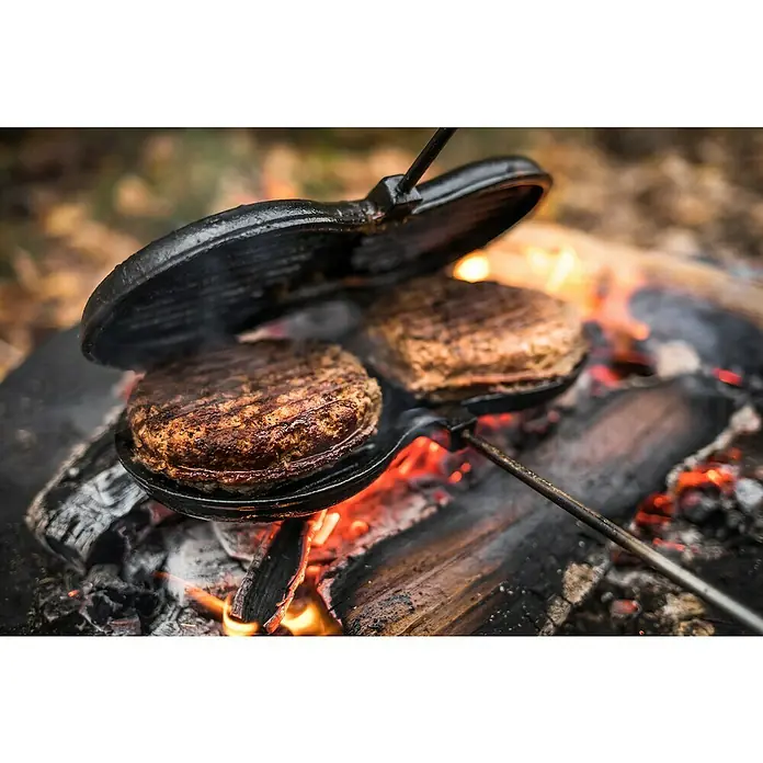 Petromax Burgereisen - L x B: 70 x 26 cm, Gusseisen - Zwei Burger-Patties werden auf einem offenen Feuer in einem gusseisernen Pie Iron zubereitet, wobei das obere Eisen aufgeklappt ist, um die Garstufe zu überprüfen.