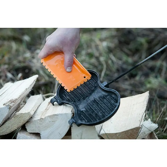 Petromax Burgereisen - L x B: 70 x 26 cm, Gusseisen - Detailaufnahme eines Petromax Stockeisen, das mit einem Schaber gereinigt wird, um seine Langlebigkeit für Outdoor-Abenteuer zu gewährleisten.