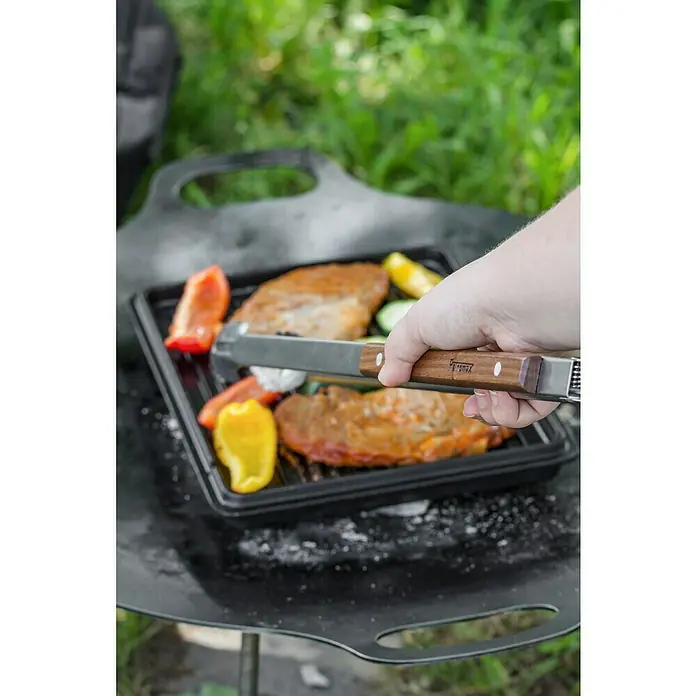 Petromax Grillzange - Länge: 41 cm, Edelstahl - Fleisch und Gemüse werden auf einer schwarzen Grillplatte im Freien gegrillt, während jemand Grillutensilien mit Holzgriffen hält.