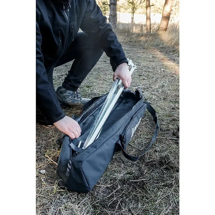 Petromax Transporttasche Dreibeinständer - Nylon, Passend für: Petromax Dreibeinständer - Eine Person verstaut ein silbernes Stativ in einer schwarzen Stativtasche, die auf dem Boden im Freien liegt. Die Tasche hat einen Tragegurt.