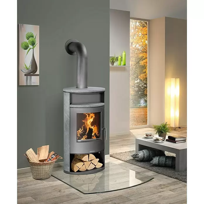 Justus Kaminofen Island Aqua - 10 kW, Raumheizvermögen: 200 m³, Material Verkleidung: Speckstein, Gussgrau - Moderner grauer Holzofen mit Glastür, die brennende Flammen zeigt, mit Holzlagerfach unten, in einem gemütlichen Wohnzimmer.