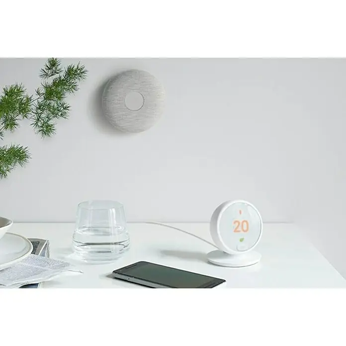 Google Nest Termostato inalámbrico inteligente - A batería, L x An x Al: 81,6 x 81,6 x 94,4 mm - Un primer plano de un termostato Nest sobre una mesa, mostrando 20 grados Celsius, junto con un teléfono inteligente y un vaso de agua.