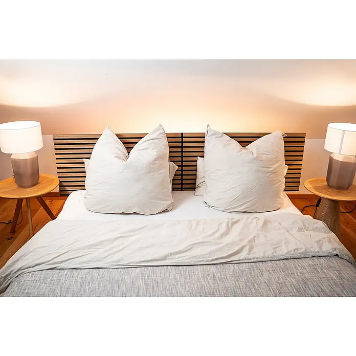 Jangal Akustikpaneel Modular Wall Furnier - Davert Oak, 1.040 x 520 x 10 mm - Einladendes Schlafzimmer mit einem modernen Bett, hervorgehoben durch übergroße Kissen und Nachttischlampen, die eine ruhige Atmosphäre schaffen.
