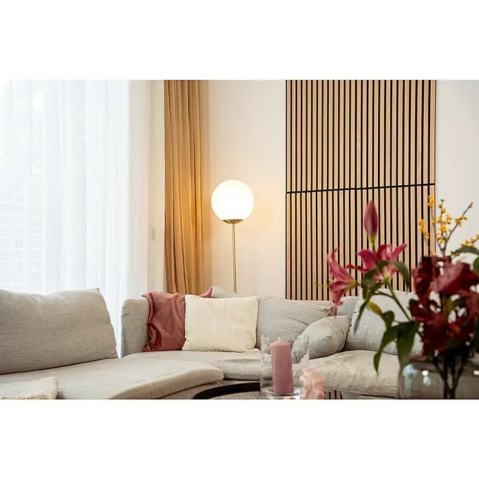 Jangal Akustikpaneel Modular Wall Furnier - Fanal Oak, 1.040 x 520 x 10 mm - Elegantes Wohnzimmer mit bequemer Couch und stilvoller Beleuchtung durch eine Stehlampe. Das Holzpaneel an der Wand ergänzt das moderne Design.