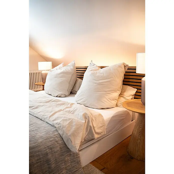Jangal Akustikpaneel Modular Wall Furnier - Davert Oak, 1.040 x 520 x 10 mm - Schlafzimmer-Interieur mit gemütlichem Bett, großen Kissen und Nachttischlampen, das eine entspannende und einladende Atmosphäre schafft.