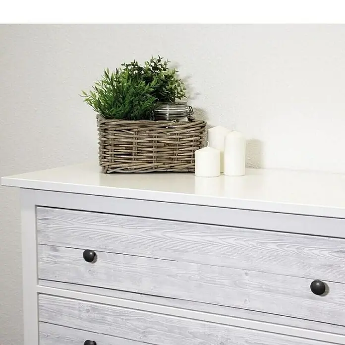 D-c-fix Holzoptikfolie Shabby Wood - 200 x 45 cm, Weiß, Selbstklebend - Nahaufnahme eines weißen Holzkommode mit dunklen Griffen, dekoriert mit einem Korb, Pflanzen und Kerzen für ein gemütliches Ambiente.