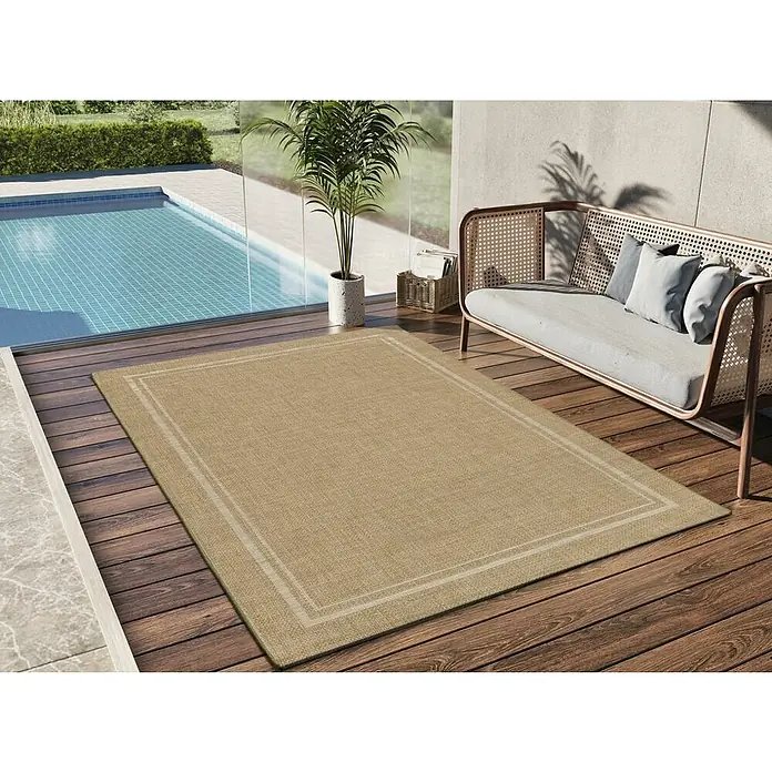 Alfombra exterior In&Out Guinea - Beige, 120 x 170 cm, Polipropileno - Una alfombra rectangular beige con borde está colocada en un patio de madera junto a una piscina y un sofá de ratán.