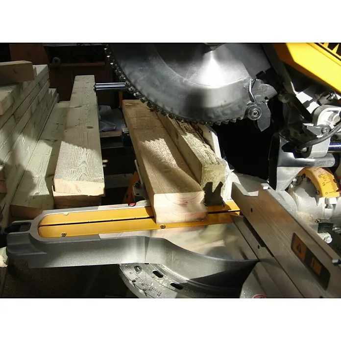 DeWalt Paneelsägen-Set DWS780KIT - 1.675 W, 0 U/min - 3.800 U/min, Durchmesser Sägeblatt: 305 mm - Eine Gehrungssäge schneidet ein Stück Holz. Die Säge hat eine gelbe und silberne Farbe und ist auf einem Werkstatttisch montiert.