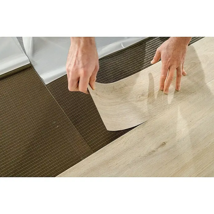 Fixed Unterlagsmatte Fixmat - 6,5 m², Stärke: 1,8 mm - Ein Mann verlegt helle, holzähnliche Vinyl-Bodenfliesen auf einem Untergrund mit Netzstruktur. Die Fliesen werden mit einer Schutzfolie versehen.
