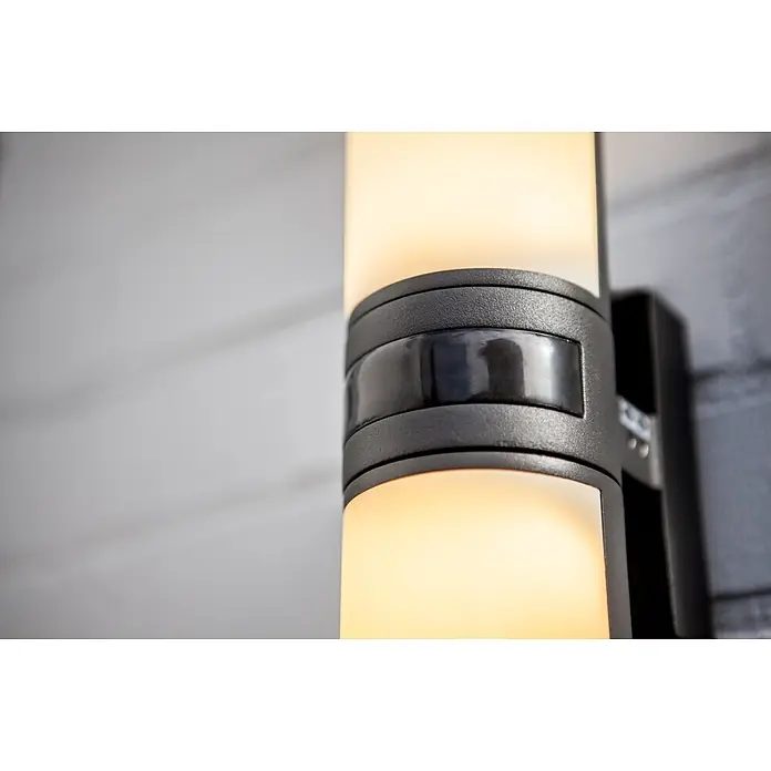 Lutec LED-Sensor-Außenwandleuchte Cyra - 16 W, L x B x H: 7,8 x 12 x 24,5 cm, Anthrazit/Weiß, Warmweiß - Nahaufnahme einer modernen, schwarzen Wandleuchte mit einem zylindrischen, diffusen Lichtkörper, der ein warmes Licht ausstrahlt.