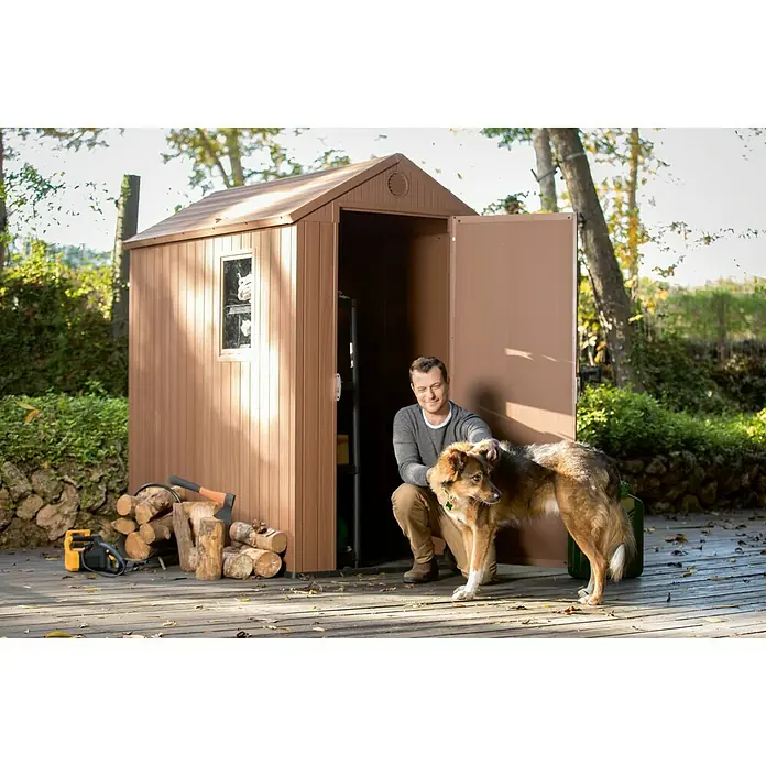 Keter Caseta de jardín Darwin 4x6 - Medidas ext. (An x Pr): 125,8 x 184,5 cm, Plástico, Marrón, 2,3 m² - Un cobertizo de jardín marrón se encuentra en una cubierta de madera, con un hombre arrodillado frente a él acariciando a un perro. Leña y una motosierra están al lado.