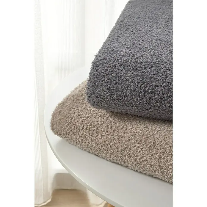 Manta para sofá Oslo - 160 x 220 cm, Poliéster, Beige - Dos mantas suaves, una gris y otra beige, están apiladas sobre una mesa redonda blanca frente a una ventana con cortinas translúcidas.