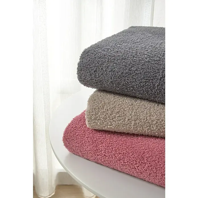 Manta para sofá Oslo - 160 x 220 cm, Poliéster, Beige - Una pila de mantas suaves en colores gris, beige y rosa, que descansan sobre una mesa blanca frente a una ventana con cortinas traslúcidas.