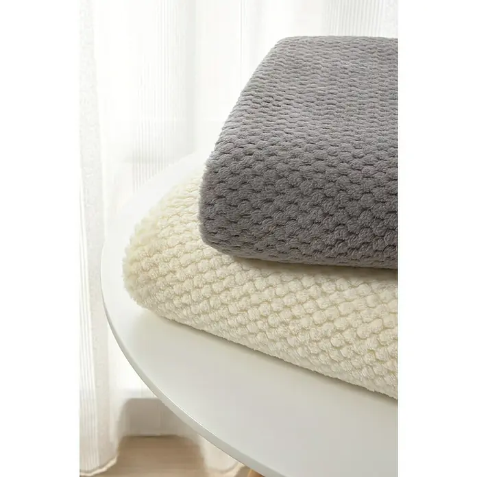 Manta para sofá Stelvio - 160 x 220 cm, Poliéster, Gris - Primer plano de dos mantas suaves y esponjosas en gris y crema, apiladas sobre una mesa blanca, con cortinas al fondo.