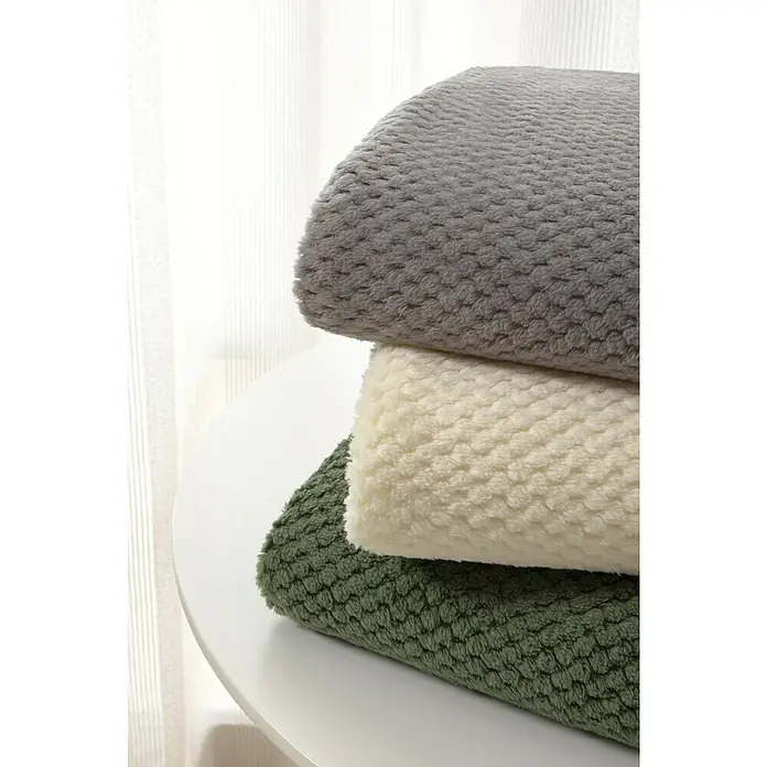 Manta para sofá Stelvio - 160 x 220 cm, Poliéster, Gris - Pila de tres mantas de felpa suaves y texturizadas en colores gris, crema y verde oliva, dispuestas sobre una mesa redonda.