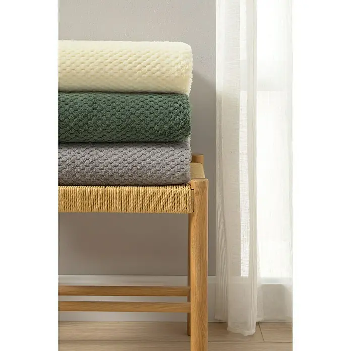 Manta para sofá Stelvio - 160 x 220 cm, Poliéster, Gris - Una pila de tres mantas suaves y afelpadas en crema, verde y gris descansa sobre un banco de madera clara.