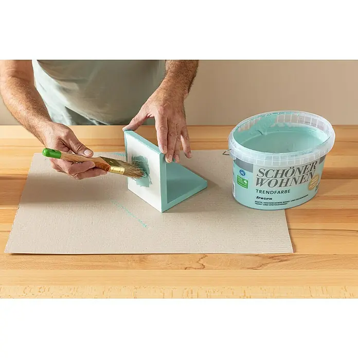 SCHÖNER WOHNEN-Farbe Wandfarbe Trendfarbe - Frozen, Matt, 1 l, Konservierungsmittelfrei - Eine Person streicht ein L-förmiges Holzstück mit einem Pinsel und aquamarinfarbener Schöner Wohnen Trendfarbe auf einer Arbeitsfläche.
