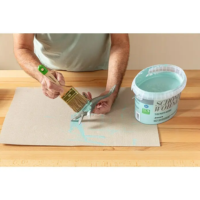 SCHÖNER WOHNEN-Farbe Wandfarbe Trendfarbe - Frozen, Matt, 1 l, Konservierungsmittelfrei - Ein Mann bemalt eine Dinosaurierfigur mit einem Pinsel und aquatischer Wandfarbe der Marke Schöner Wohnen.