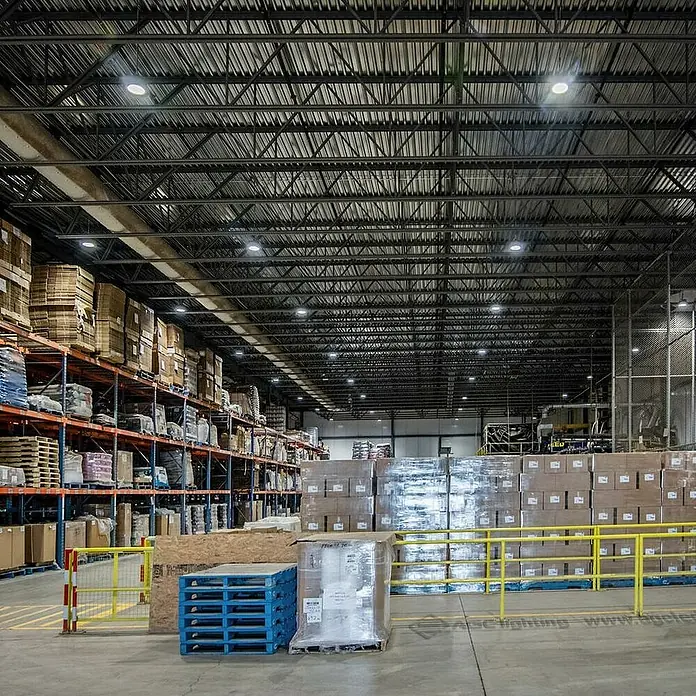 Led Hispania Campana LED Merak-II - 100 W, Diámetro: 24,4 cm, Aluminio, 3.000 K - 6.000 K - Un almacén iluminado con estanterías altas llenas de mercancías y paletas aseguradas con flejes y envoltura de lámina.