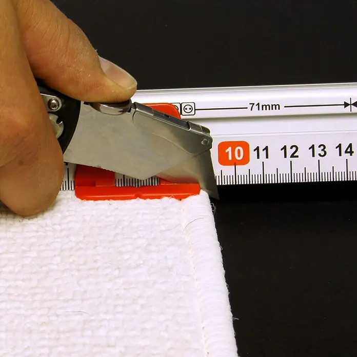 Connex Regla de marcado - Largo: 60 cm, Aluminio - Primer plano de un cúter para alfombras con carril metálico que se utiliza para recortar una alfombra blanca al tamaño deseado.