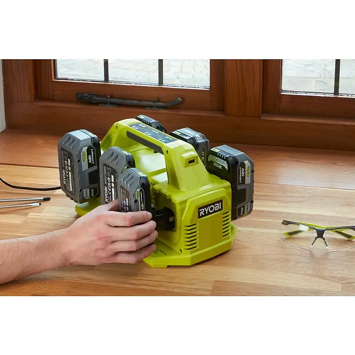 Ryobi ONE+ Akku-Ladegerät RC18640 - Passend für: Ryobi One+ 18V Akkus - Ein gelbes Ryobi Ladegerät mit mehreren Akkus, das auf einer Holzoberfläche steht, wobei eine Hand einen Akku einsetzt.