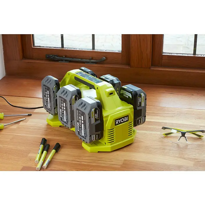 Ryobi ONE+ Akku-Ladegerät RC18640 - Passend für: Ryobi One+ 18V Akkus - Ein gelbes Ryobi-Ladegerät mit mehreren Akkus, flankiert von Schraubendrehern, Markern und einer Schutzbrille auf einem Holztisch.