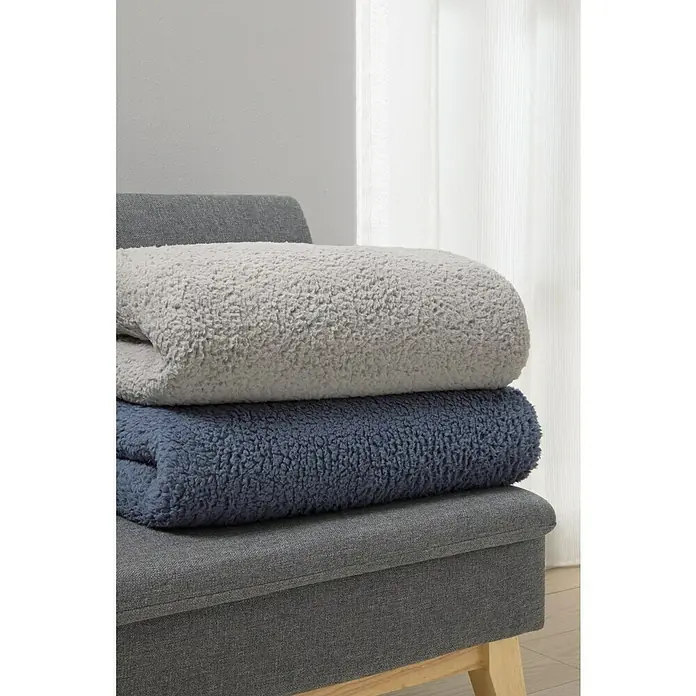 Manta para sofá Lucerna - 170 x 230 cm, Poliéster, Gris - Una pila de dos mantas Sherpa acogedoras en tonos de gris y azul, colocadas sobre un otomano gris, invitan a la relajación.