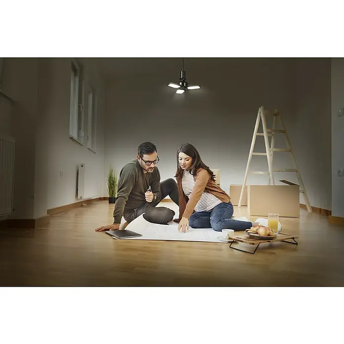 Osram Deckenleuchte Highbay E27 Blade - 40 W, L x B x H: 33,7 x 33,7 x 10,9 cm, Schwarz, E27 - Ein Paar plant in einem neuen Haus, im Fokus steht eine moderne Deckenleuchte. Sie sitzen auf dem Boden, umgeben von Umzugskartons und Bauplänen.