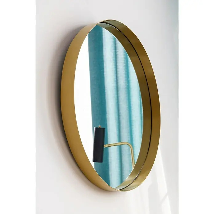 Espejo de pared Gia con marco - Diámetro: 60 cm, Oro - Primer plano de un espejo redondo dorado en una pared blanca, que refleja una lámpara y cortinas azules en la habitación.