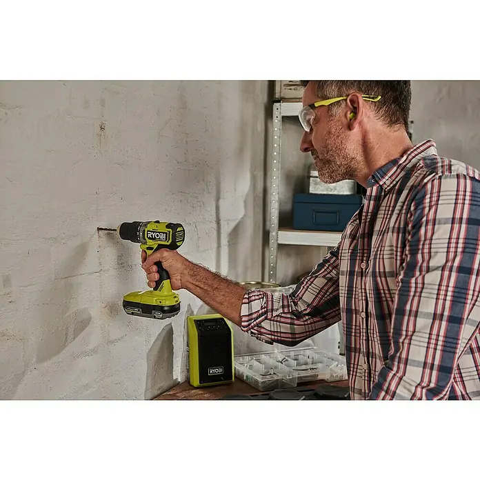 Ryobi ONE+ Akku-Schlagbohrschrauber RPD18C1-0 C - 18 V, Ohne Akku, 60 Nm - Ein Mann bohrt mit einem Ryobi Akku-Bohrschrauber ein Loch in eine weiße Wand. Er trägt Schutzbrille und Gehörschutz für seine Sicherheit.