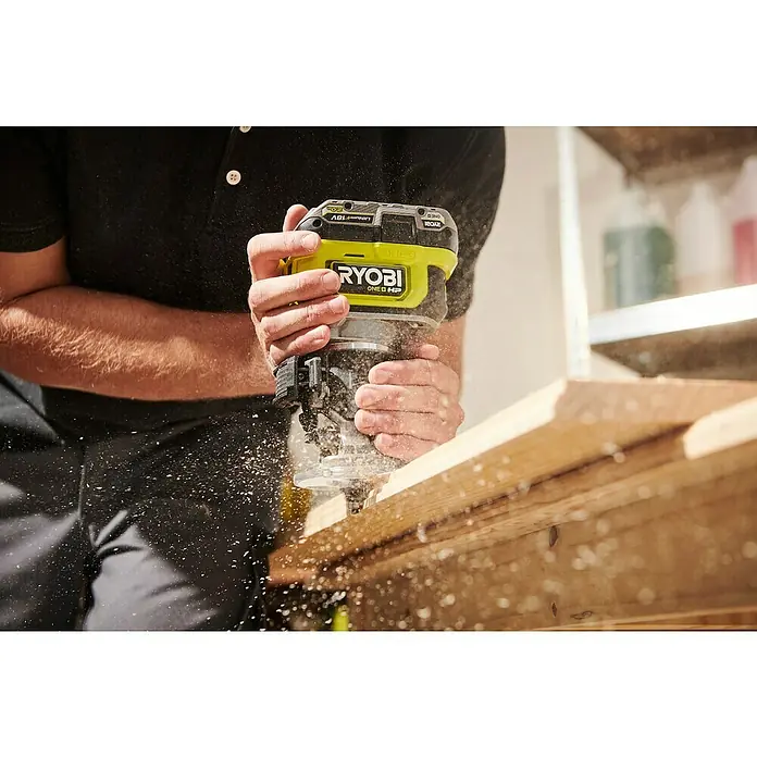Ryobi ONE+HP Akku-Kantenfräser RTR18C-0 - 18 V, Ohne Akku - Nahaufnahme von Händen, die eine RYOBI ONE+ HP Kompakt-Oberfräse beim Bearbeiten eines Holzstücks halten, wobei Holzspäne herumfliegen.