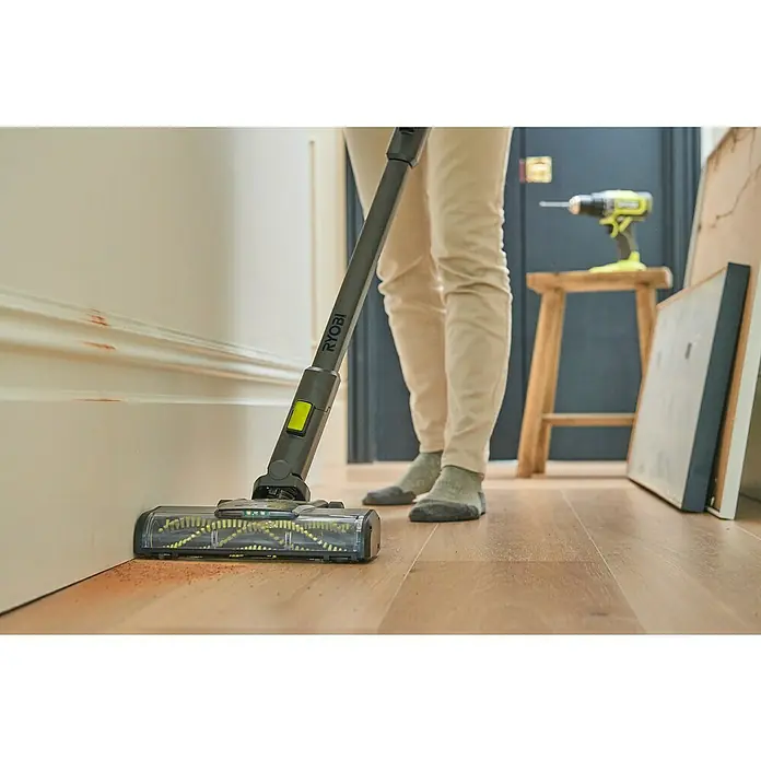 Ryobi ONE+ Akku-Staubsauger RSV18BL - 18 V, 1 Akku, 4 Ah, Behältervolumen: 0,8 l - Ein Ryobi-Staubsauger wird verwendet, um den Boden entlang einer weißen Wand zu reinigen. Auf einem Holzhocker steht ein Ryobi-Bohrer.